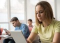 High school student reading a lesson from digital tablet while her classmates are doing the same thing in the background.